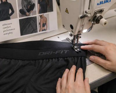 A stretch-knit waistband with a Dri-Fit label being sewn on an industrial machine.