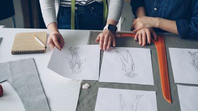 Two people reviewing pattern sketches on a worktable, marking them with a ruler.